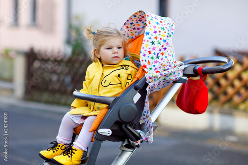 Portrait of little cute toddler girl sitting in stroller or pram and going for a walk. Happy cute baby child having fun outdoors. Healthy daughter. Street traffic in the city.