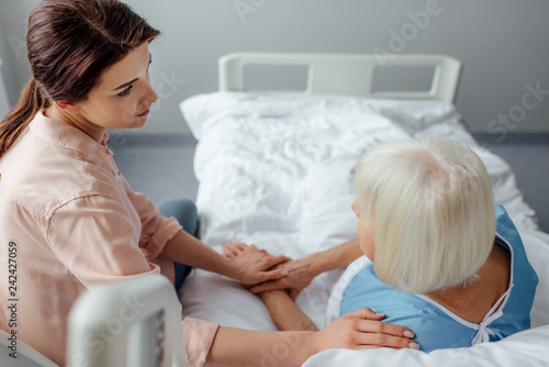 selective focus of senior woman and upset daughter holding hands in hospital