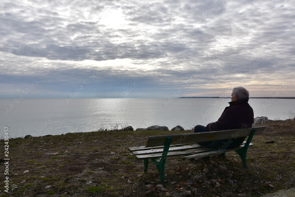 Silhouette of a man over 50 years sitting on an old wooden bench above the see in a cloudy winter day. Senior man quietly watching the see. Nostalgic mood. Duran Kulak, Bulgaria