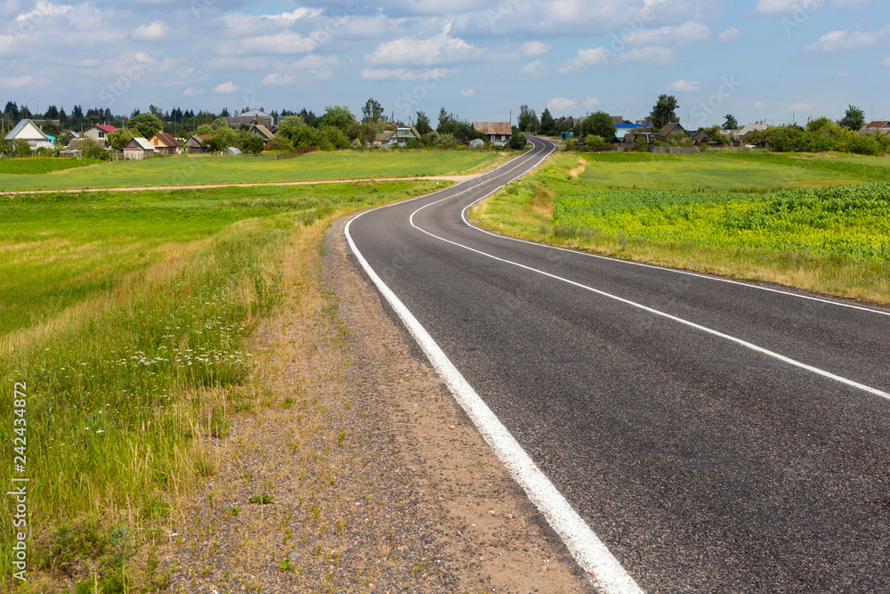 View of the small asphalt road in the countryside with blue sky and cumulus clouds on the background.