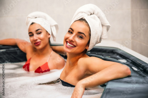 Young european and asian women sit together in jacuzzi. They wear swimsuits and towels around hair. Cheerful models smile on camera. Water is full of foam.