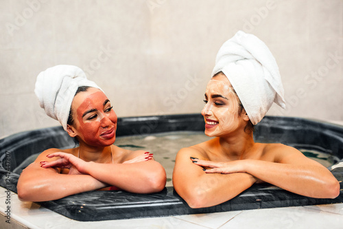 Delightful and cheerful young women relaxing in jacuzzi. Theu keep hands crossed and look at each other. Models smile. They have mask on face and towels on hair.