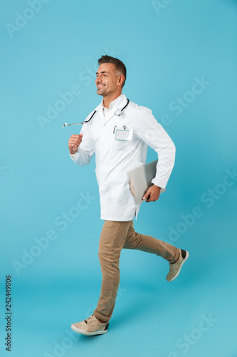 Full length image of joyous man wearing white medical coat and stethoscope running, while isolated over blue background photo