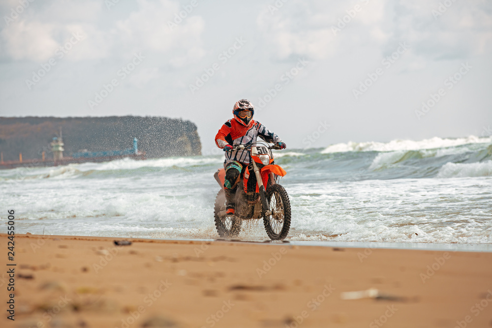 Obraz premium Motorcyclist in a protective suit rides a motorcycle on the sea, splashes fly from under the wheels.