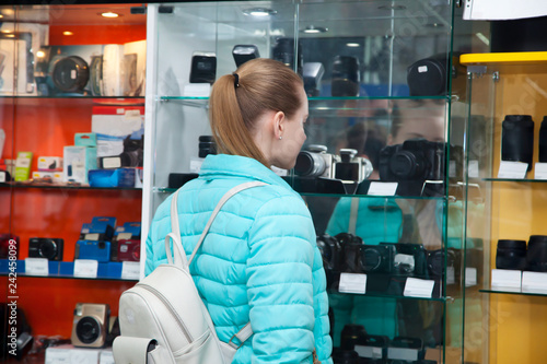 Young woman standing near showcase with photo cameras. View from the back