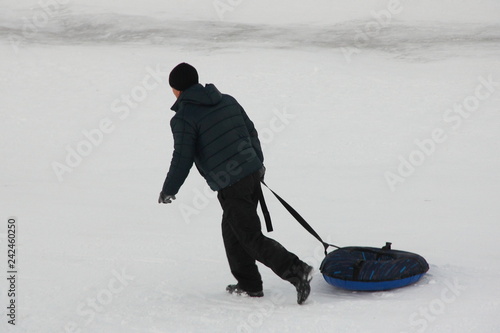 Men in black jacket and hat flee with inflatable snowtubing on snow at winter day - side rear view