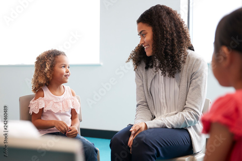 Infant school girl and her female teacher sitting on chairs in a classroom smiling to each other, close up