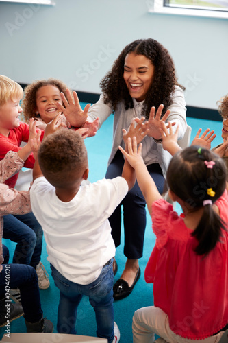 Elevated view of infant school children in a circle in the classroom giving high fives to their smiling female teacher, vertical, close up