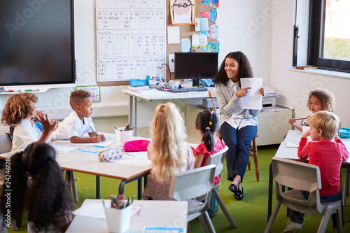 Принти на полотні Smiling female infant school teacher sitting on a chair facing school kids in a