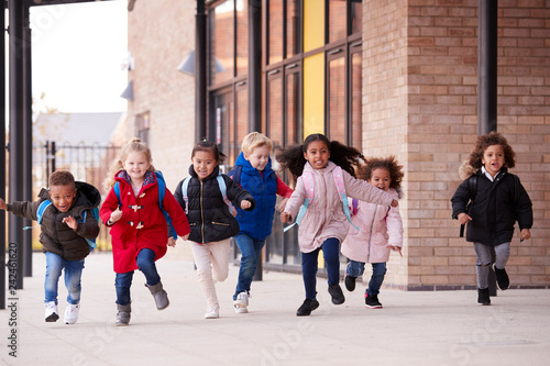 A happy multi-ethnic group of young school kids wearing coats and carrying schoolbags running in a walkway with their classmates outside their infant school building photo