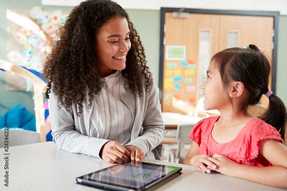 Female infant school teacher working one on one with a Chinese schoolgirl, sitting at a table in a classroom using a tablet computer, looking at each other, close up Female infant school teacher working one on one with a Chinese schoolgirl, sitting at a table in a classroom using a tablet computer, looking at each other, close up