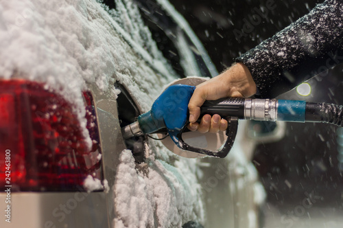 A man in a black coat fills his car at a gas station on a snowy winter evening. Hand holding a blue refueling gun..