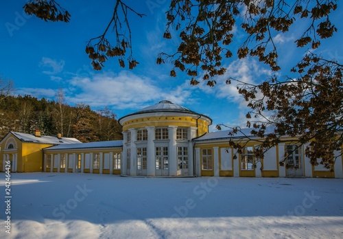 Yellow building of Ferdinand colonnade with mineral water at spa town Marienbad, Czech Republic, snow winter scene in park, shadows on ground, sunny day, blue sky, white clouds, branch with dry leaves