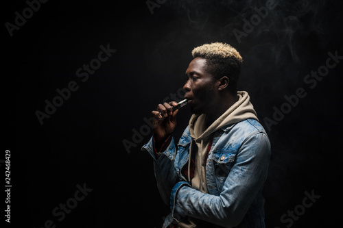 African Man smoking or vaping e-cig or electronic cigarette holding a mod with a lot of clouds isolated on black background