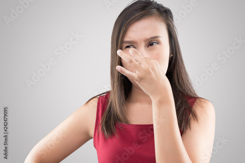Asian woman in red shirt holding her nose because of a bad smell on gray background.