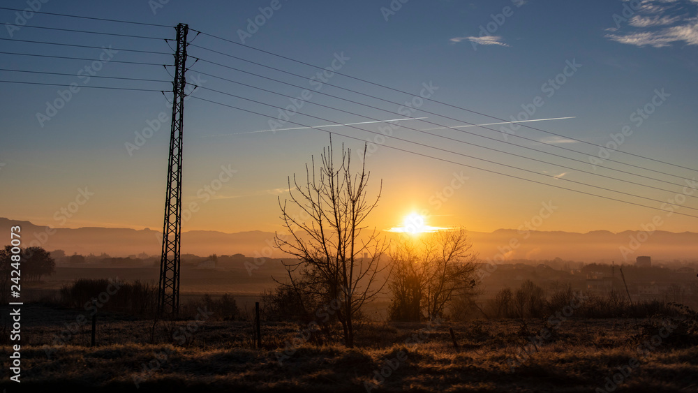 Sun rising on a dark cloudscape with electricity towers on a blue and yellow morning scene on a plain plant landscape