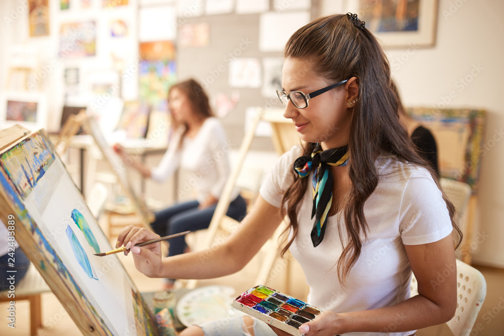 Fototapeta premium Beautiful brown-haired girl in glasses dressed in white t-shirt and jeans with a scarf around her neck paints a picture in the art studio