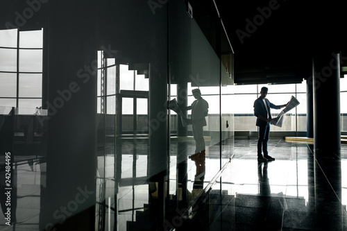 Architect in stylish clothes holds sheet with drawing in his hand and talks by phone on the background of a modern glass multistory building