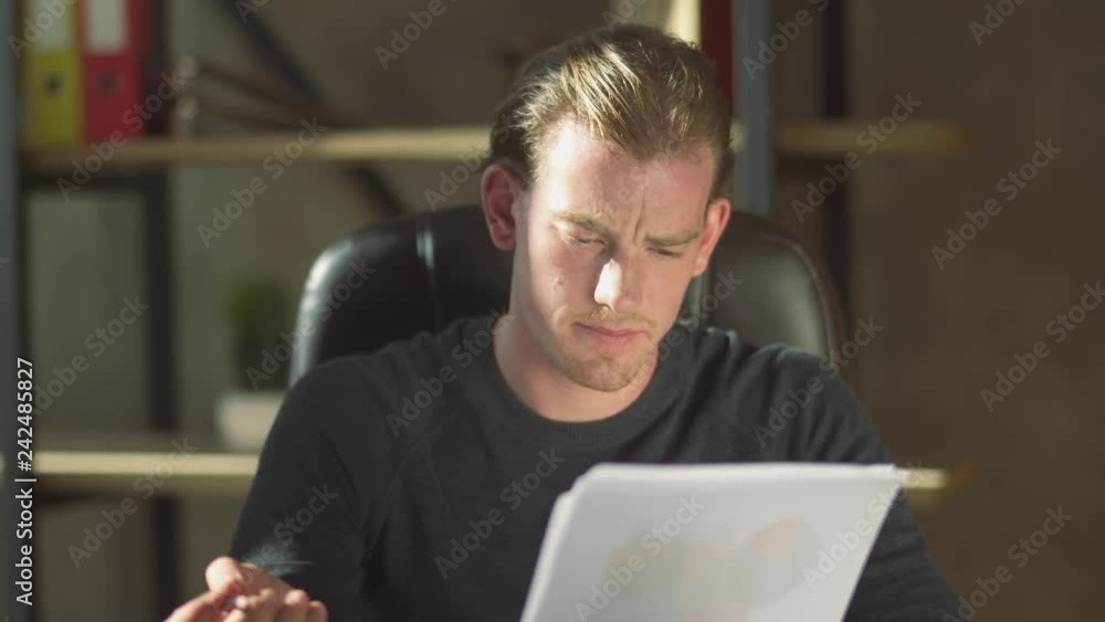 Young thoughtful puzzled businessman sitting in the office at the table near notebook and working at documents, make amendment in papers. Young thoughtful puzzled businessman sitting in the office at the table near notebook and working at documents, make amendment in papers.