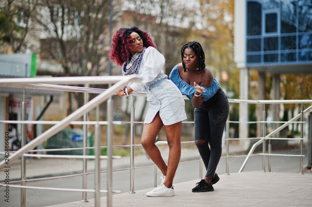 Obraz premium Two attractive african american womans posed near railings against modern multistory building. Hit on buttocks.