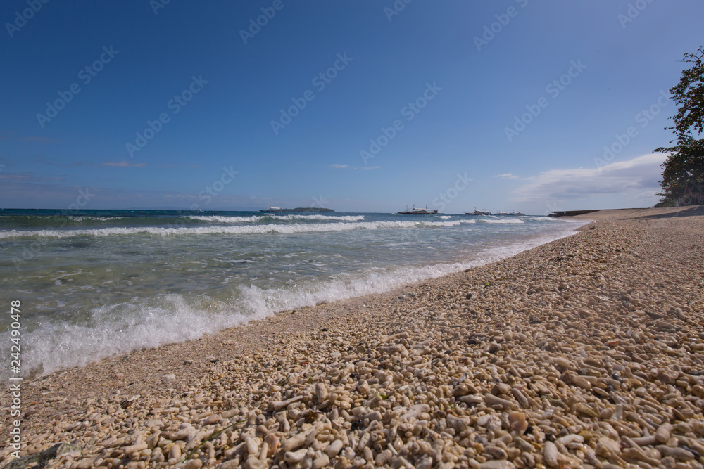 beach with coral stones pebbles at the seashore with waves and boats in the background on a day with blue sky and white clouds  