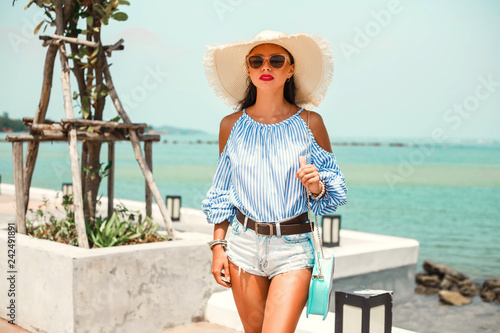 Beautiful lady in a striped T-shirt and denim shorts  sunglasses and a straw hat. Positive emotions. Walking through the shopping center  the concept of summer holidays sea and shopping