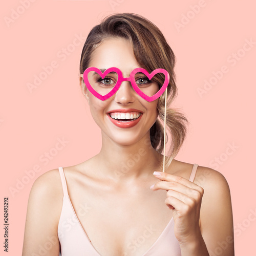 Portrait of cheerful smiling woman holding paper party glasses heart in hand. Pink background.