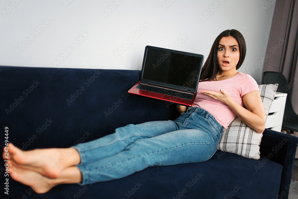 Naklejka premium Upset and distracted young woman sit on sofa in room. She hold red laptop. Young woman point on black screen. Laptop is broken. It doesn't work.