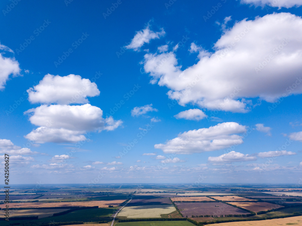 Panorama from the air on the lush meadows and fields with colorful plots of agricultural crops grown under the open sky outside the city in the sun under a blue sky and white clouds. Panorama from the air on the lush meadows and fields with colorful plots of agricultural crops grown under the open sky outside the city in the sun under a blue sky and white clouds.