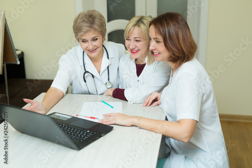 three women doctor in white coats sit at the table in the clinic in the office with a laptop