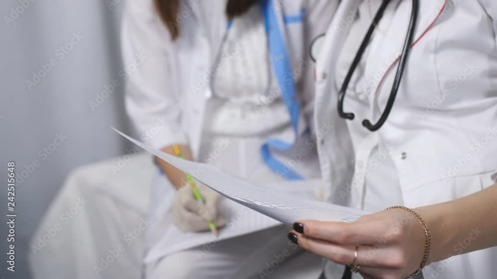 Two beautiful young doctors women double check files with paper documents. Two women doctor in the examining room filing papers, close-up. Two beautiful young doctors women double check files with paper documents. Two women doctor in the examining room filing papers, close-up.