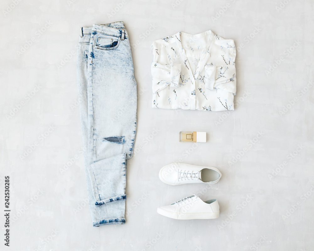 Blue jeans, white shirt with floral print, white sneakers and perfume lying  on grey background. Overhead view of woman's casual day outfits. Trendy  hipster look. Top view of women's clothes. Stock Photo