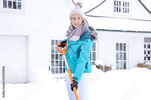 hübsche junge blonde frau schippt im winter den weißen Schnee vor ihrem haus auf einen grossen haufen - mit kälteschutz blauer jacke und mütze bekleidet lächelt sie glücklich photo