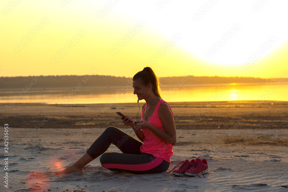 Young beautiful girl in sportswear sits on a sandy beach and enjoys an orange sunset by the sea and listens to music in her mobile phone