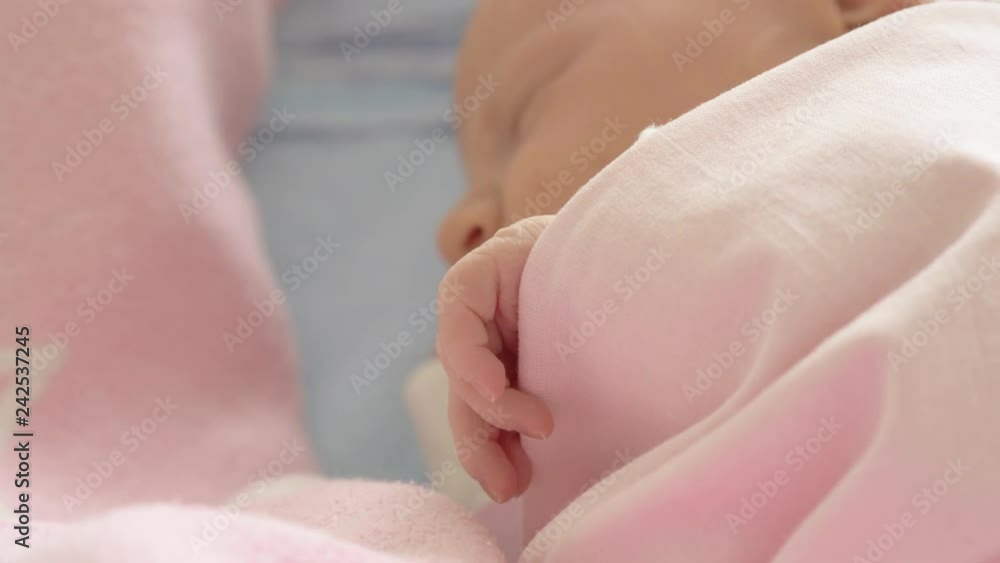 A closeup of a newborn girls face, who is sleeping under the cozy pink blanket. Her hand is out of it and tiny cute fingers are holding the edge of soft fabric A closeup of a newborn girls face, who is sleeping under the cozy pink blanket. Her hand is out of it and tiny cute fingers are holding the edge of soft fabric