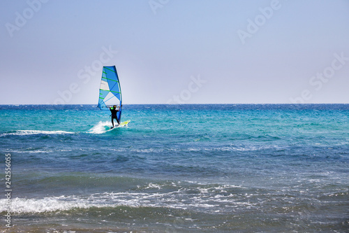 Surfer Spending Time on Outdoor Water Sport Adventure. Clear Sky and Blue Wave. Person Balancing on a Windsurfing Board with Sail. Vacation Activity ideas