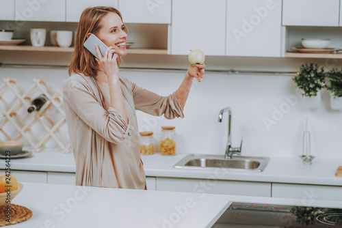 Cheerful lady with apple is speaking on phone in kitchen