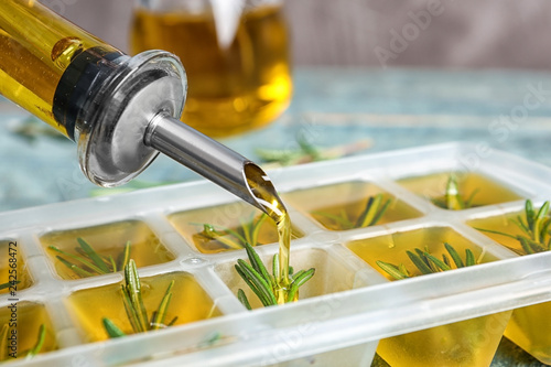 Pouring olive oil into ice cube tray with rosemary on table, closeup