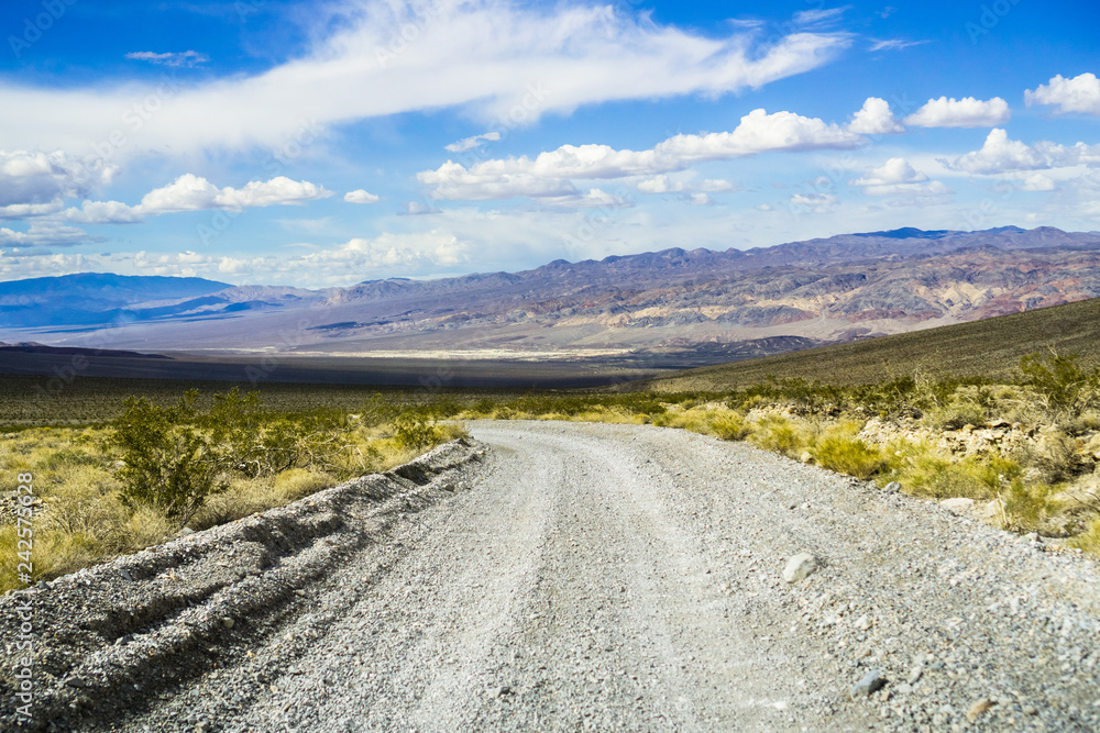 Travelling on an unpaved road through a remote area of Death Valley National Park; mountains, blue sky and white clouds in the background; California