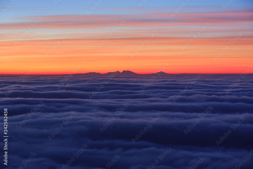 Abstract artistic background image - Sunrise above the clouds, sea of clouds with mountain silhouette in the distance. Beautiful vibrant red and yellow clouds above the horizon, tranquility in nature