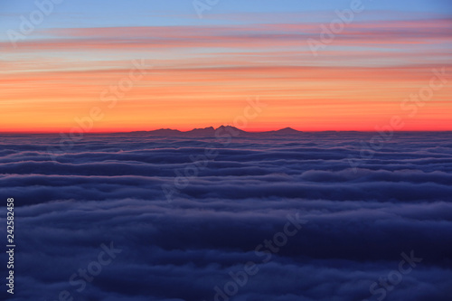 Abstract artistic background image - Sunrise above the clouds, sea of clouds with mountain silhouette in the distance. Beautiful vibrant red and yellow clouds above the horizon, tranquility in nature