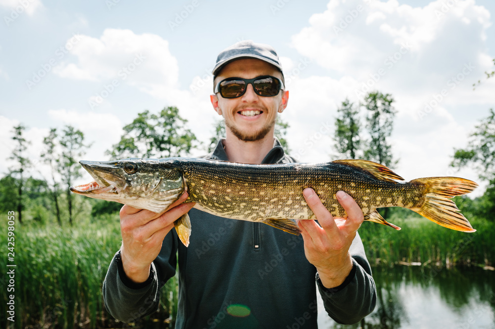 Happy cheerful young fisherman hold a big fish pike on a background of lake and nature. Fishing background. Good catch. Trophy fish. angler. headshot. close up. Happy cheerful young fisherman hold a big fish pike on a background of lake and nature. Fishing background. Good catch. Trophy fish. angler. headshot. close up.