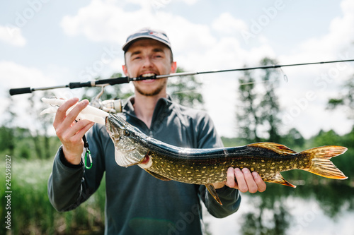 Happy cheerful young fisherman hold a big fish pike on a background of lake and nature. Fishing background. Good catch. Trophy fish. angler. headshot. close up.