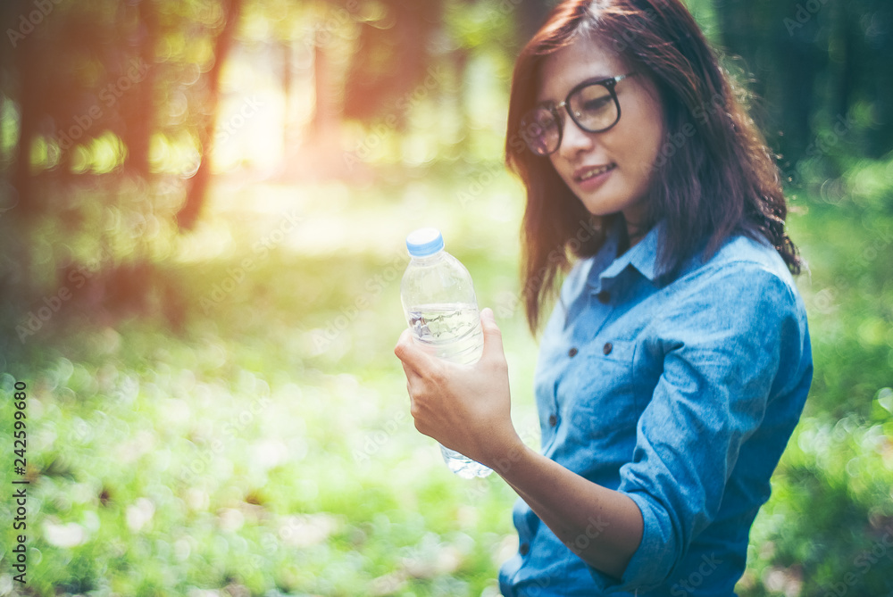 In hot weather, strong sun and a tropical forest. Hiking, beautiful Asian girl holding her bottle of purified water bottles and water bottles handed out ahead with a brilliant smile with copy space. In hot weather, strong sun and a tropical forest. Hiking, beautiful Asian girl holding her bottle of purified water bottles and water bottles handed out ahead with a brilliant smile with copy space.