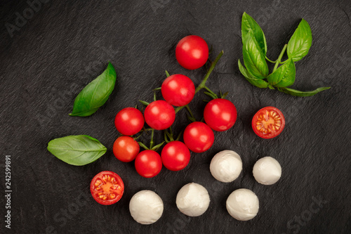An overhead photo of Mozzarella cheese, cherry tomatoes and fresh basil leaves on a black background, Italian cuisine ingredients with copyspace