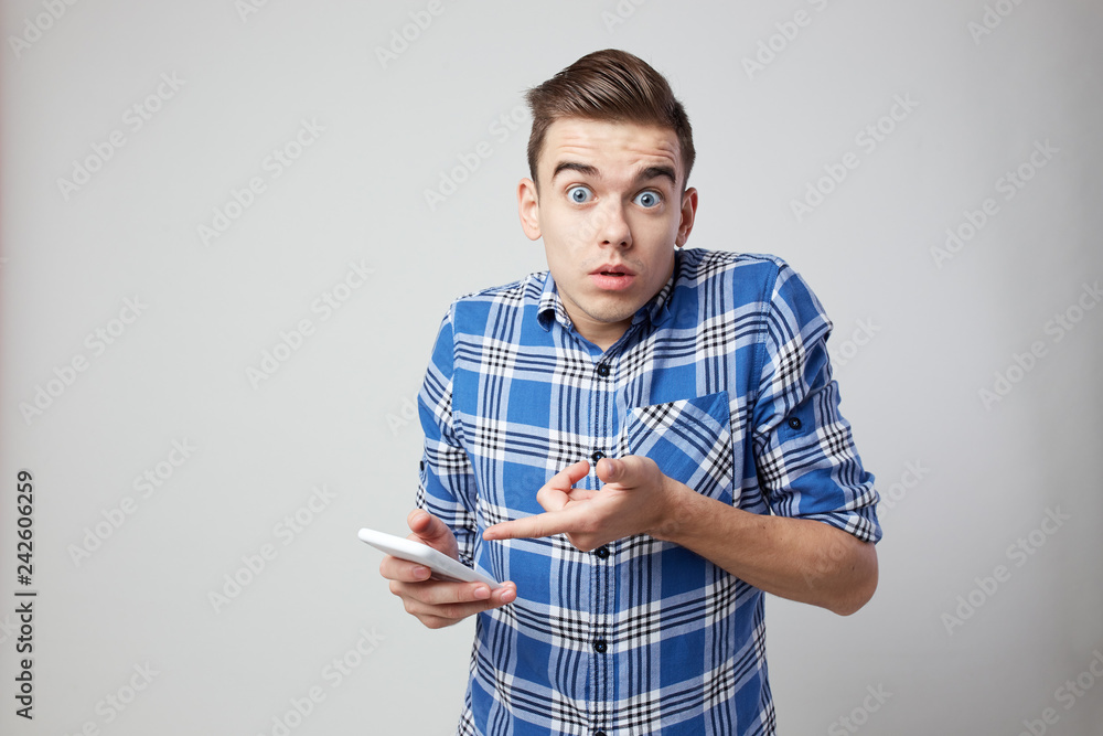 Surprised guy dressed in a plaid shirt and jeans keeps mobile phone in his hand on a white background in the studio Surprised guy dressed in a plaid shirt and jeans keeps mobile phone in his hand on a white background in the studio
