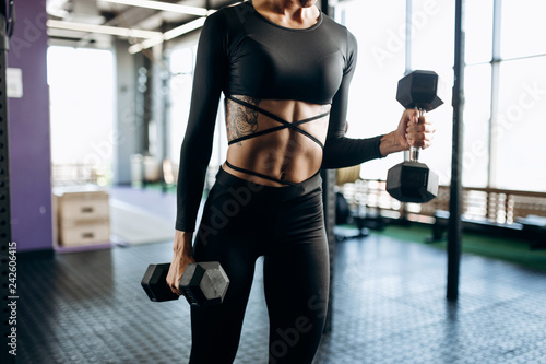 Slim body of a young woman with tattoo in a black sportswear that builds up muscles with dumbbells in the gym