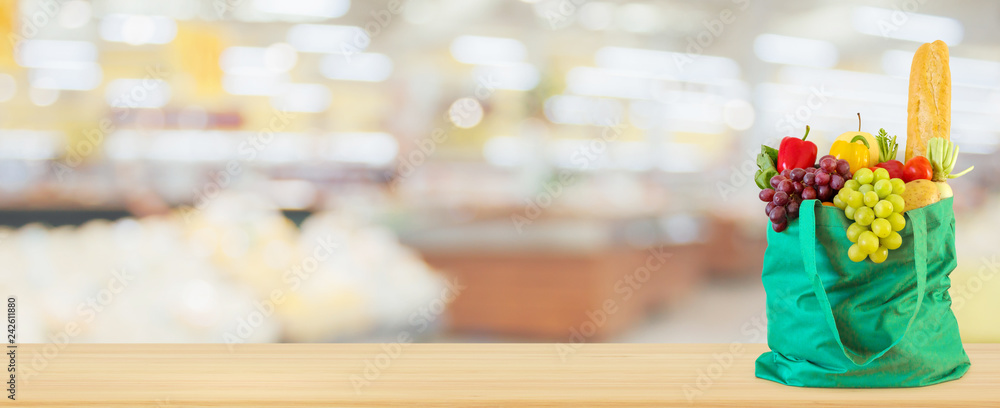 Fresh fruits and vegetables in reusable green shopping bag on wood table top with supermarket grocery store blurred defocused background with bokeh light