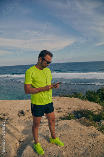 Jogger / runner holding a cellphone with earbuds outdoors with ocean / sea background.