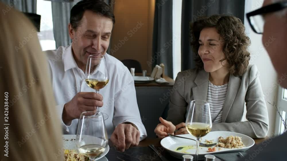 Joyous middle aged businessman holding wine glass, smiling and giving a toast while having lunch with colleagues in the restaurant